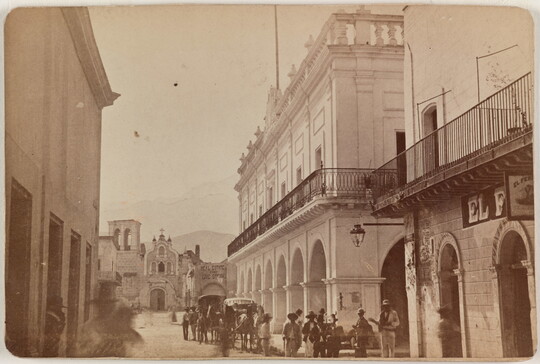 A sepia-toned photograph of a street scene with tall buildings on either side, people and carriages in the street, mountains in the distance.