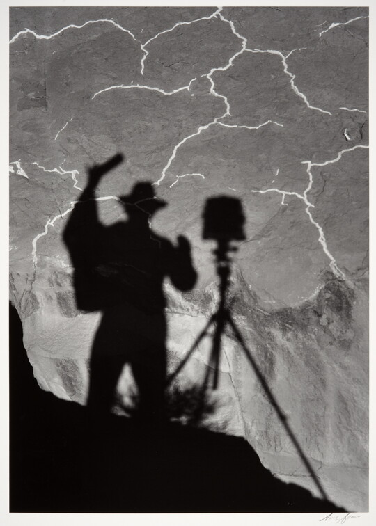 A black-and-white photograph of the shadow of a person and tripod with camera cast from above onto the landscape below.