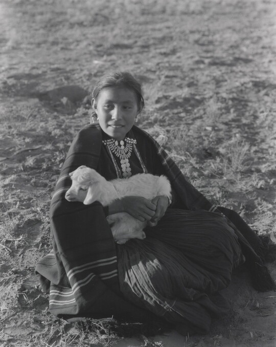A black-and-white photograph of an Indigenous girl sitting on the ground while holding a lamb.