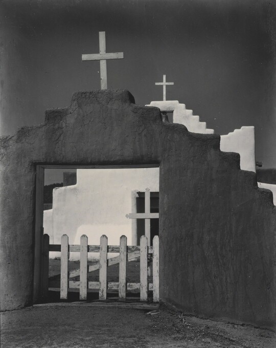 A black-and-white photograph of a white adobe church seen through the entrance of the surrounding adobe wall topped with a wooden cross.