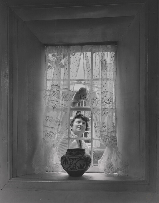 A black-and-white photograph of a sheer curtained window with a Native American-style pot on the sill as a White woman cleans the window from outside.