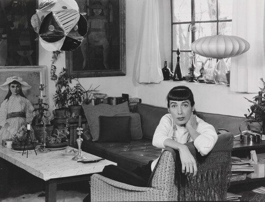 A black-and-white photograph of a White woman sitting sideways in a chair, facing the camera, in a room full of art, sculpture, bottles, candlesticks, etc.