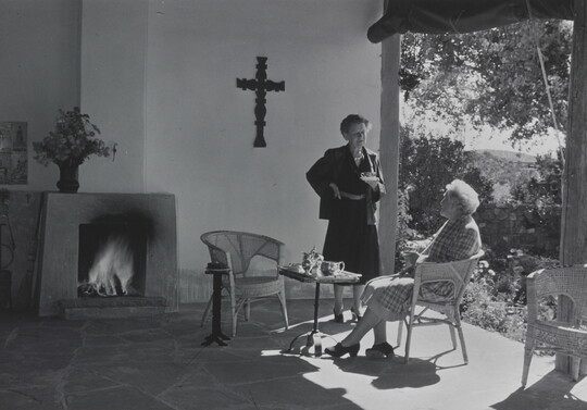 A black-and-white photograph of two White women, one seated and one standing, talking on a covered patio.