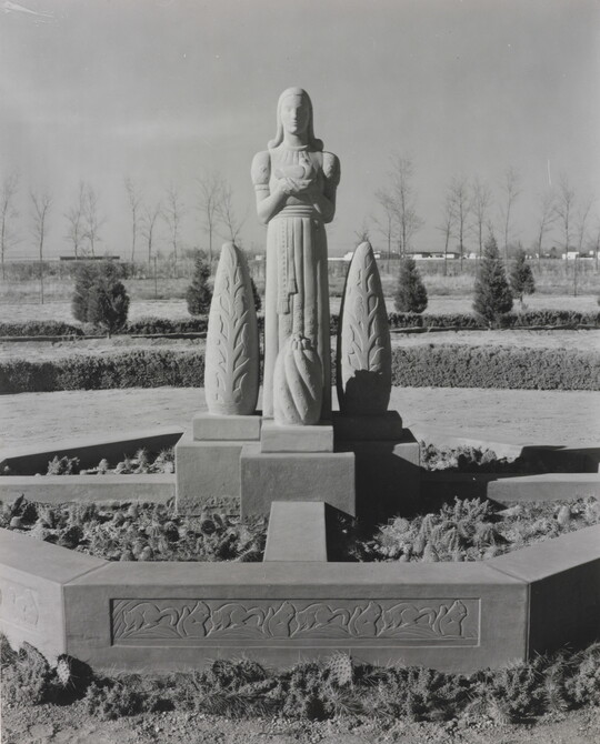 A black-and-white photograph of a stone statue of a stylized figure standing between stylized stone trees.