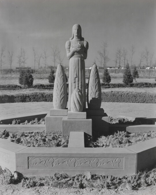 A black-and-white photograph of a stone statue of a stylized figure standing between stylized stone trees.