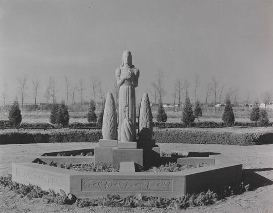 A black-and-white photograph of a stone statue of a stylized figure standing between stylized stone trees.
