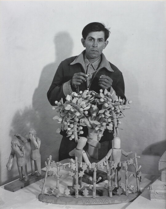 A black-and-white photograph of a medium-skinned man standing behind a wood sculpture of trees and human figures.