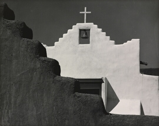 A black-and-white photograph of a terraced adobe bell tower with a cross on top, partially hidden by a terraced adobe wall.