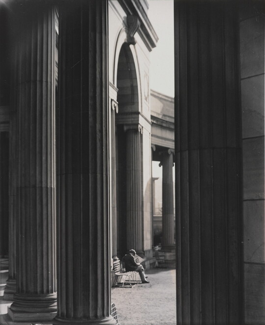 A black-and-white photograph of a person sitting on a bench reading a newspaper as seen from between large fluted columns.