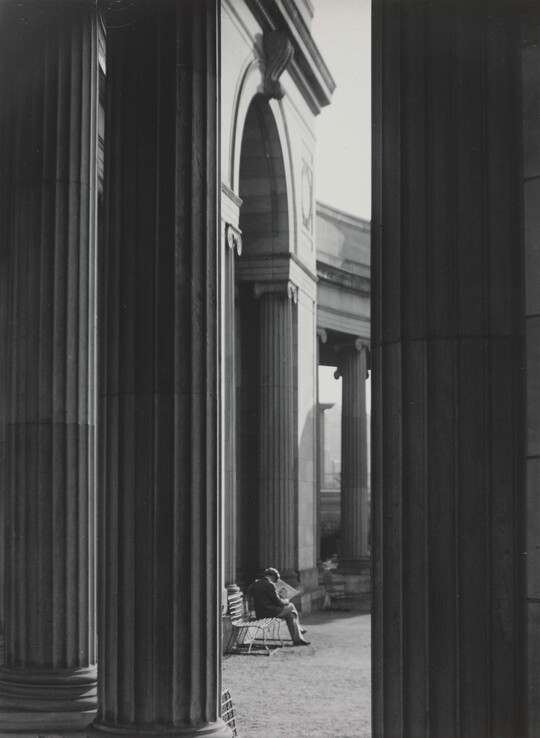 A black-and-white photograph of a person sitting on a bench reading a newspaper as seen from between large fluted columns.