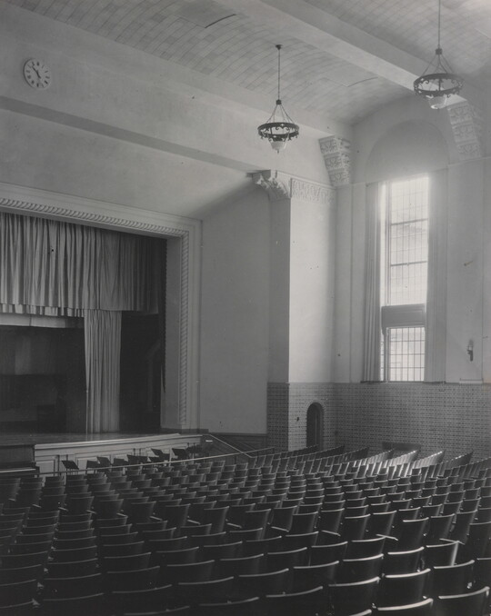 A black-and-white photograph of an empty auditorium showing rows of seats, a stage, and a tall window.