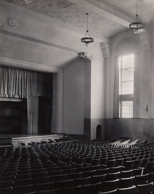 A black-and-white photograph of an empty auditorium showing rows of seats, a stage, and a tall window.