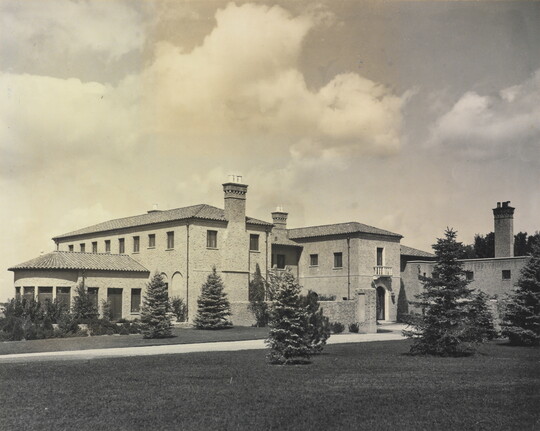 A black-and-white photograph of a large multi-story mansion with a mission-style roof and a large grass yard with evergreens.