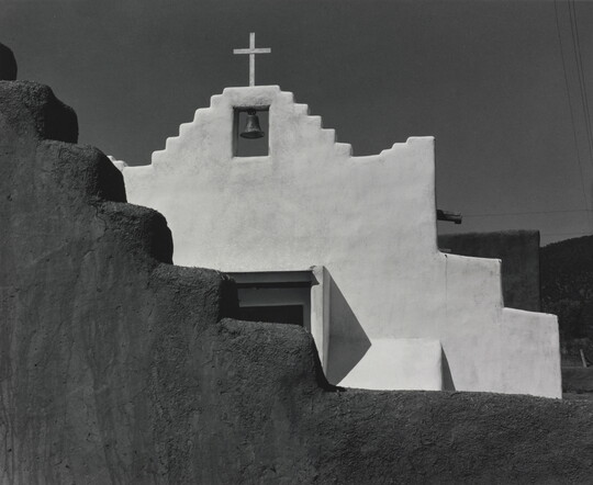 A black-and-white photograph of a terraced adobe bell tower with a cross on top, partially hidden by a terraced adobe wall.