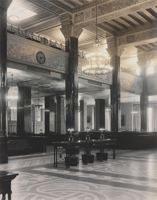 A black-and-white photograph of the interior of an ornate office lobby with inlaid floors, columns, and chandeliers.