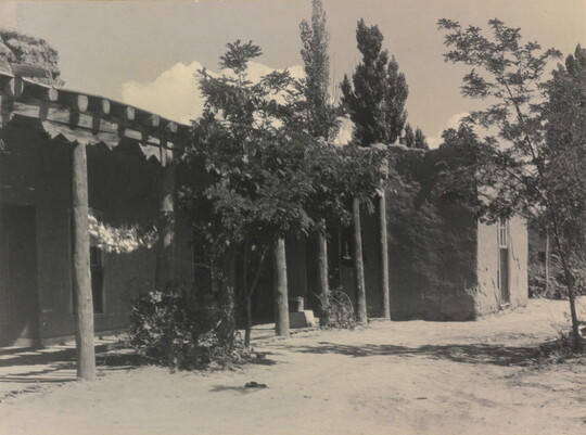 A black-and-white photograph of the exterior of a single-story adobe house and covered porch surrounded by trees and a dirt yard.