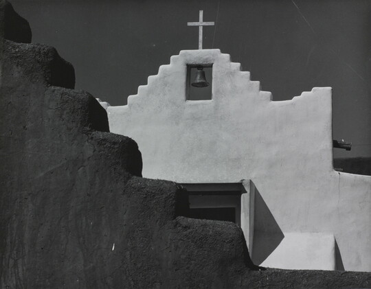 A black-and-white photograph of a terraced adobe bell tower with a cross on top, partially hidden by a terraced adobe wall.