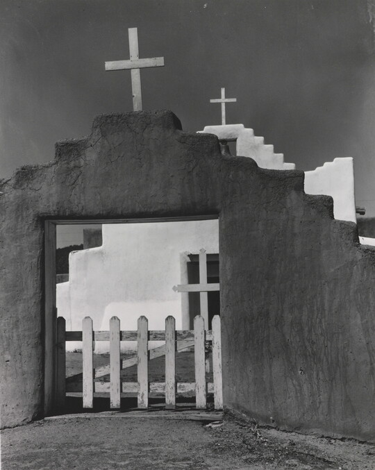 A black-and-white photograph of a white adobe church seen through the entrance of the surrounding adobe wall topped with a wooden cross.