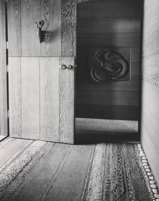 A black-and-white photograph of a Dutch door open to a decorative wood and stone floor and a carved sculpture on the far wall.