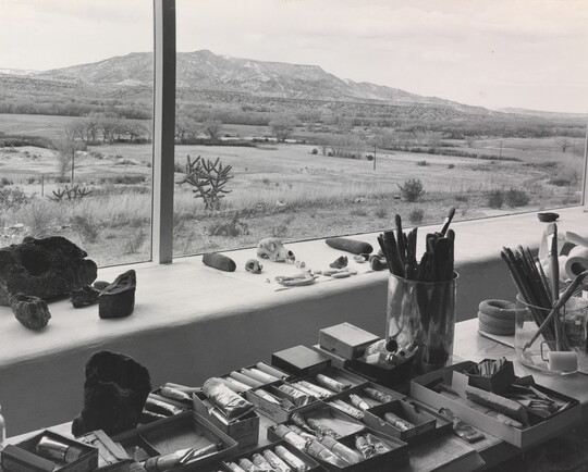 A black-and-white photograph of paints, brushes, and other art supplies on a wide windowsill that overlooks a mountain landscape.