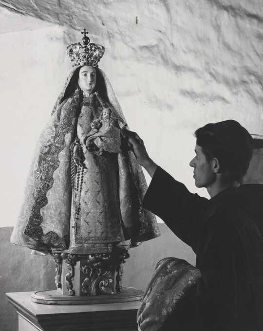 A black-and-white photograph of a friar touching a statue of the Virgin Mary dressed as queen of heaven holding the infant Jesus.