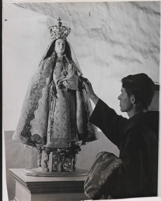 A black-and-white photograph of a friar touching a statue of the Virgin Mary dressed as queen of heaven holding the infant Jesus.