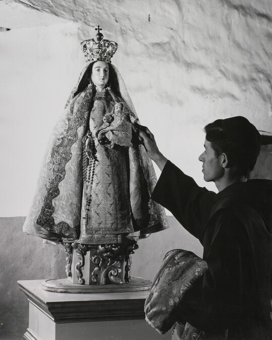 A black-and-white photograph of a friar touching a statue of the Virgin Mary dressed as queen of heaven holding the infant Jesus.
