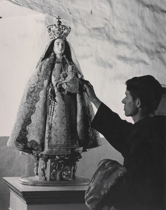 A black-and-white photograph of a friar touching a statue of the Virgin Mary dressed as queen of heaven holding the infant Jesus.