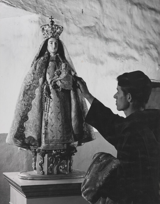 A black-and-white photograph of a friar touching a statue of the Virgin Mary dressed as queen of heaven holding the infant Jesus.