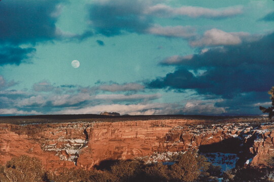 A color photograph of an almost-full moon in a cloudy blue sky over red, rocky cliffs with snow in the crevices.