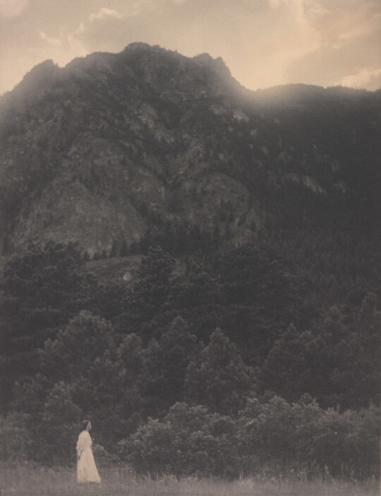 A black-and-white photograph of a woman in a white dress standing at the base of a rocky mountain covered with trees.
