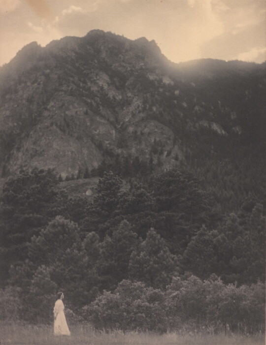A black-and-white photograph of a woman in a white dress standing at the base of a rocky mountain covered with trees.