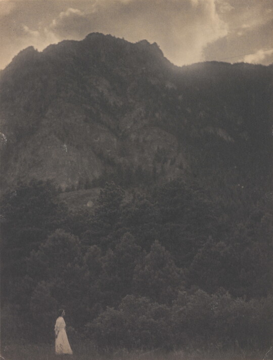 A black-and-white photograph of a woman in a white dress standing at the base of a rocky mountain covered with trees.