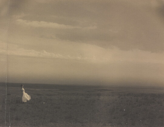 A black-and-white photograph of a woman in a white dress standing in a large meadow.