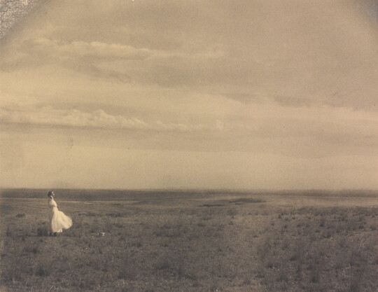 A black-and-white photograph of a woman in a white dress standing in a large meadow.