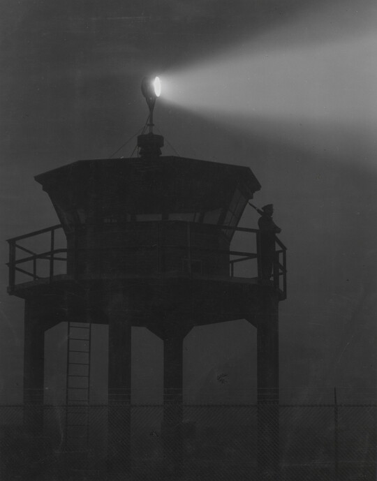 A black-and-white photograph of a person standing in silhouette atop a tower with a bright spotlight shining from the top.