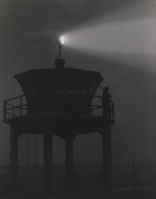 A black-and-white photograph of a person standing in silhouette atop a tower with a bright spotlight shining from the top.