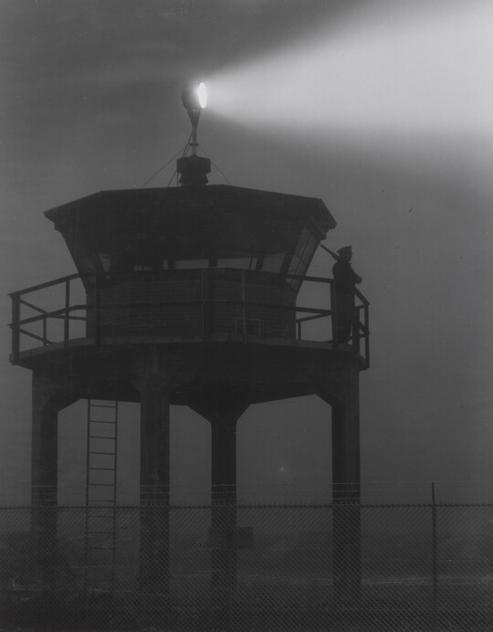 A black-and-white photograph of a person standing in silhouette atop a tower with a bright spotlight shining from the top.