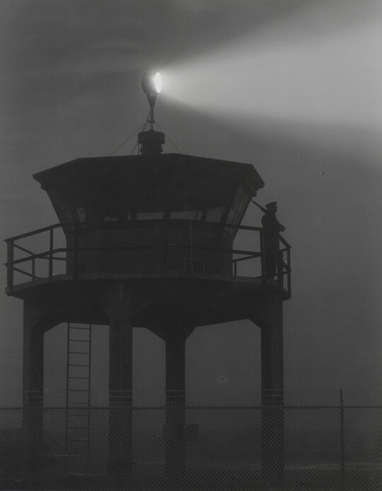 A black-and-white photograph of a person standing in silhouette atop a tower with a bright spotlight shining from the top.