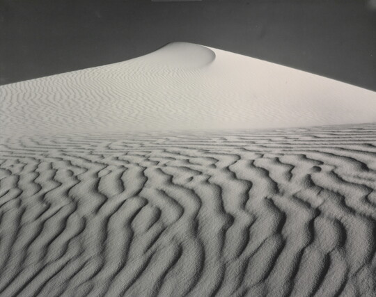 A sepia-toned photograph of a deeply-rippled sand dune rising to a point into the sky.