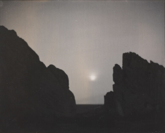 A black-and-white photograph of a moon shining through a foggy sky from between the V-shaped silhouette of large rocks.