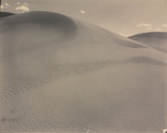A sepia-toned photograph of a rippling sand dune rising up into a sky dotted with a few small clouds.