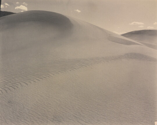 A sepia-toned photograph of a rippling sand dune rising up into a sky dotted with a few small clouds.