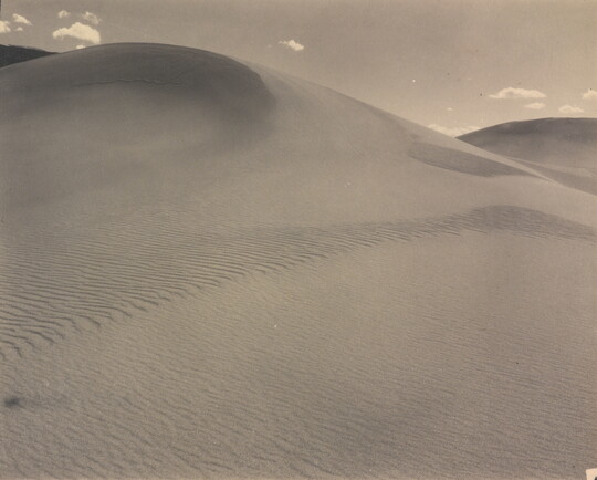 A sepia-toned photograph of a rippling sand dune rising up into a sky dotted with a few small clouds.