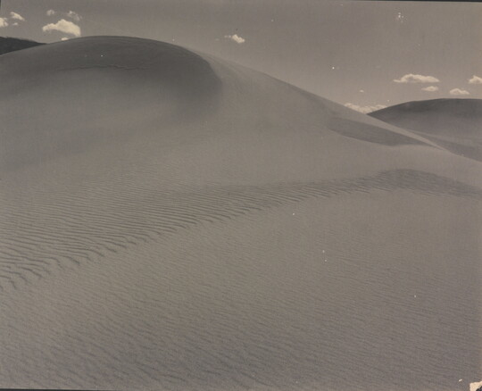 A sepia-toned photograph of a rippling sand dune rising up into a sky dotted with a few small clouds.