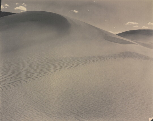 A sepia-toned photograph of a rippling sand dune rising up into a sky dotted with a few small clouds.