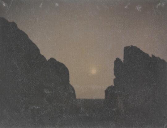 A black-and-white photograph of two rock formations in silhouette with a hazy moon in between them.