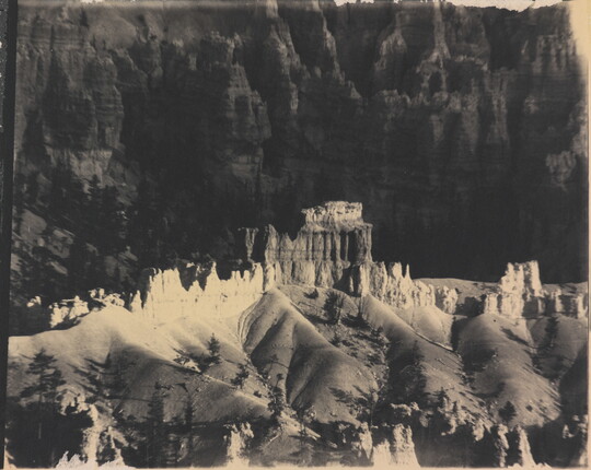 A black-and-white photograph of a landscape with hoodoos, or vertical spires of rock, on top of sand dune shapes with cliffs in the background.