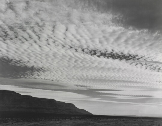 A black-and-white photograph of rippling white clouds over a flat plain with a mountain rising on the left side.