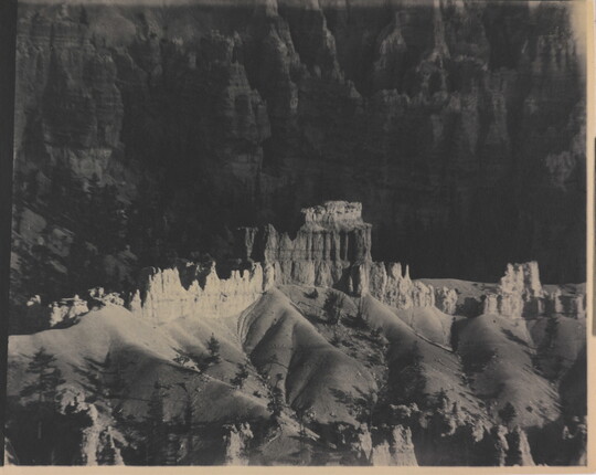 A black-and-white photograph of a landscape with hoodoos, or vertical spires of rock, on top of sand dune shapes with cliffs in the background.